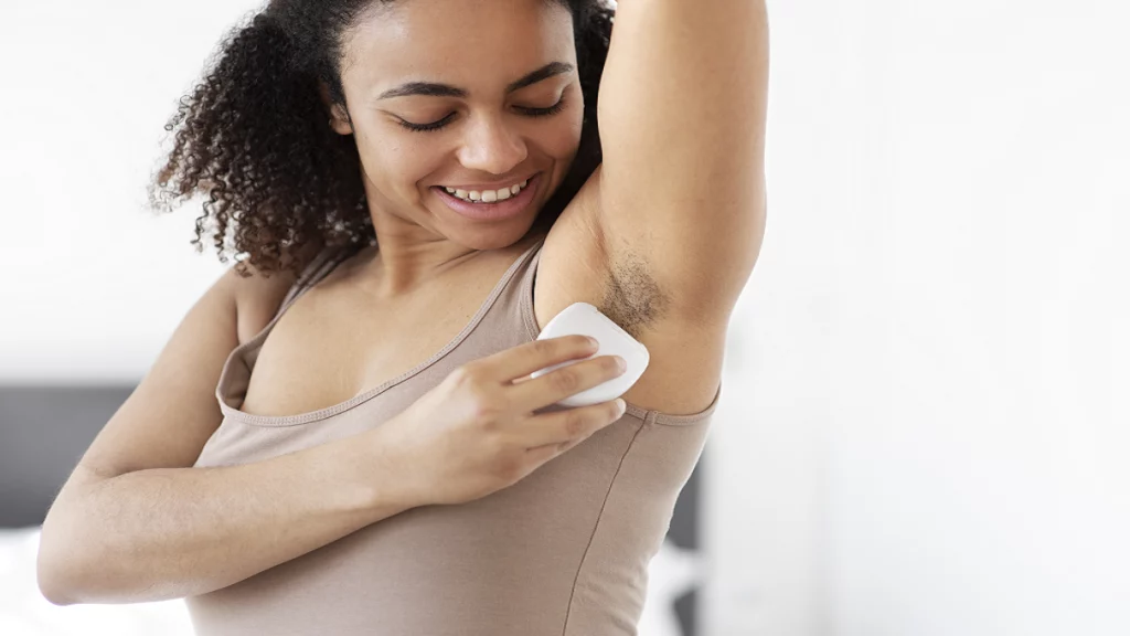 A imagem mostra uma mulher aplicando desodorante em suas axilas. Ela sorri e olha para essa região enquanto faz a aplicação.