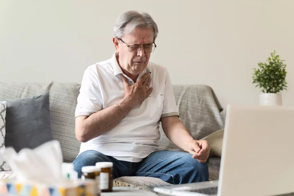 Na imagem é possível ver um homem branco idoso, que veste uma camisa polo branca, sentado em um sofá na frente do notebook com a mão sobre o peito.