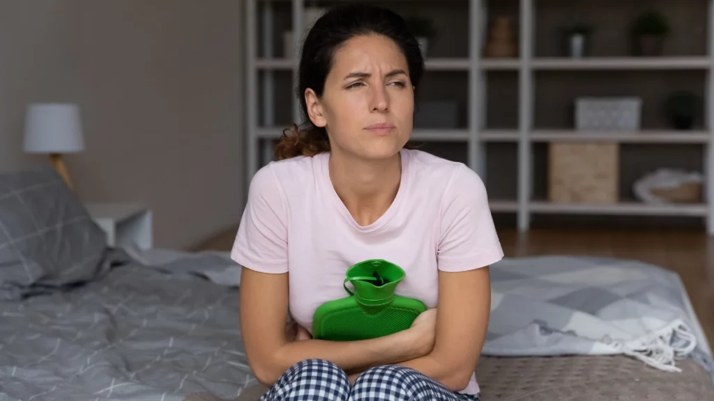 dr.consulta - Mulher com cólica por cisto no ovário. Ela está sentada na cama, segurando bolsa térmica na bariga.
