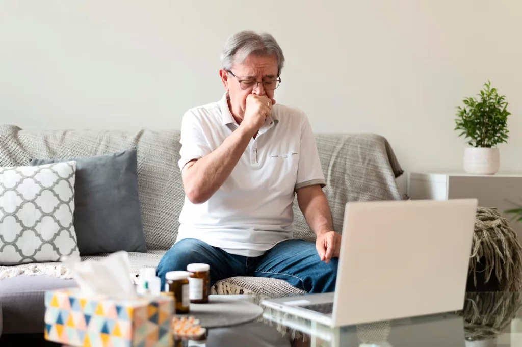 A imagem mostra um senhor sentado em seu sofá na frente de um computador. Ele está com a mão na região da boca e aparenta sentir dor ao engolir.