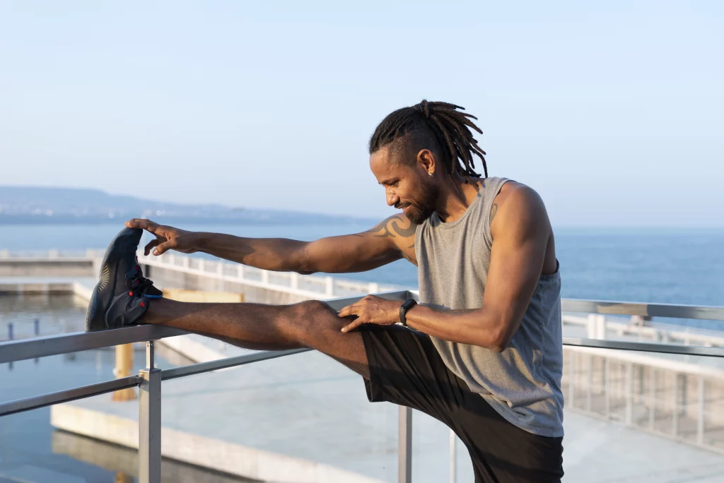 Na imagem há um homem negro com dreads no cabelo alongando uma de suas pernas em uma barra de metal. Ele veste shorts preto, camiseta regata cinza e tênis pretos.