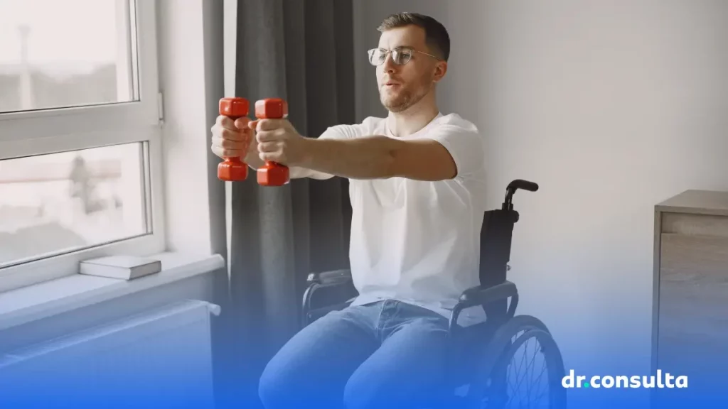 Na imagem é possível ver um homem branco em cadeira de rodas fazendo exercícios de braço em um quarto. Ele está levantando pequenos halteres vermelhos.