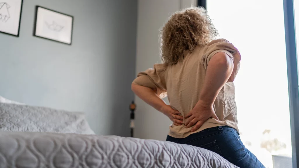 dr.consulta - Mulher sentada na cama com as mãos nas costas sinalizando lombalgia, um tipo de doenças reumáticas.
