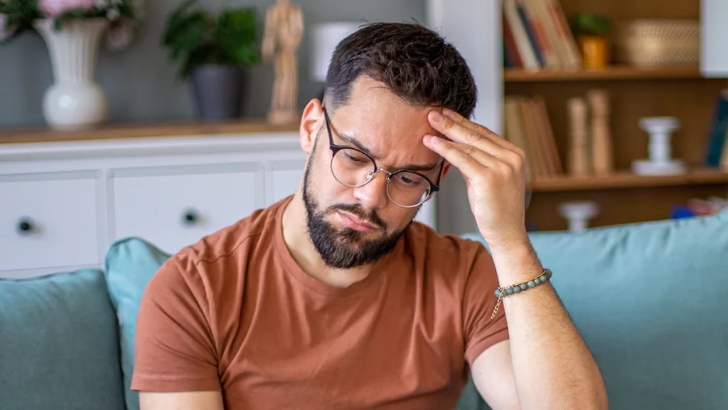 dr.consulta - Homem jovem com expressão de preocupação sentado em um sofá, segurando a cabeça, ilustrando sintomas e desafios da esclerose múltipla. 
