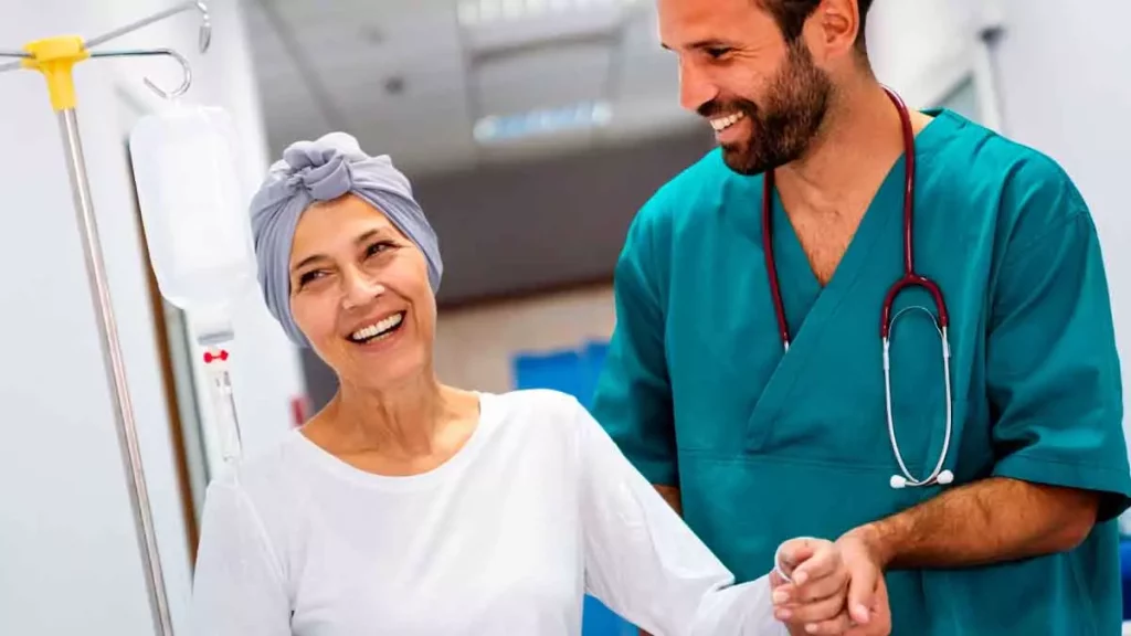 Mulher mais velha portadora de leucemia crônica ao lado do seu médico. Uma das mãos estão dadas e ambos estão sorrindo.