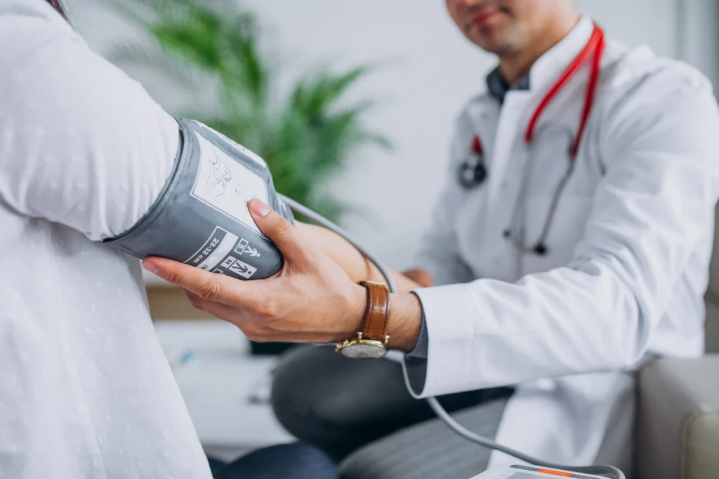 A imagem foca na mão de um médico que está aferindo a pressão arterial de uma paciente, ela veste uma camisa branca.