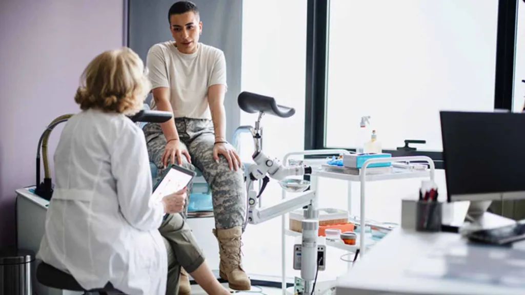 Mulher branca sentada em uma maca de hospital e vestindo camiseta branca e calça militar conversando com uma médica durante uma consulta. O rosto da médica não está visível. Também existem equipamentos médicos ao fundo.