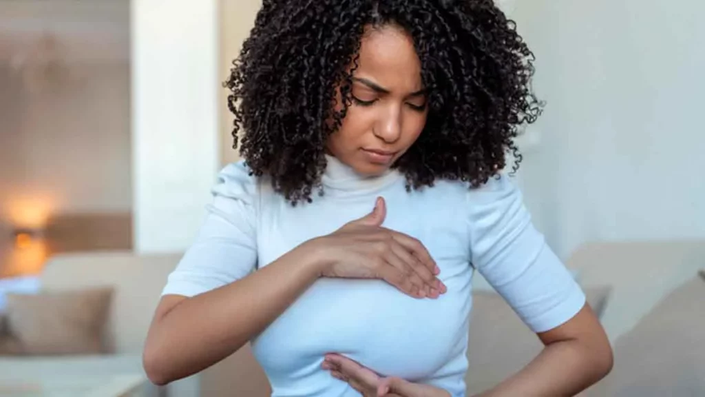 Imagem de uma mulher negra realizando o autoexame de mama. Ela está com uma blusa branca e apalpando seu seio esquerdo.