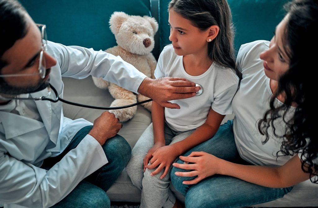 Profissional pediatra utilizando o estetoscópio em uma garota. No consultório também há uma mulher abraçando a menina.