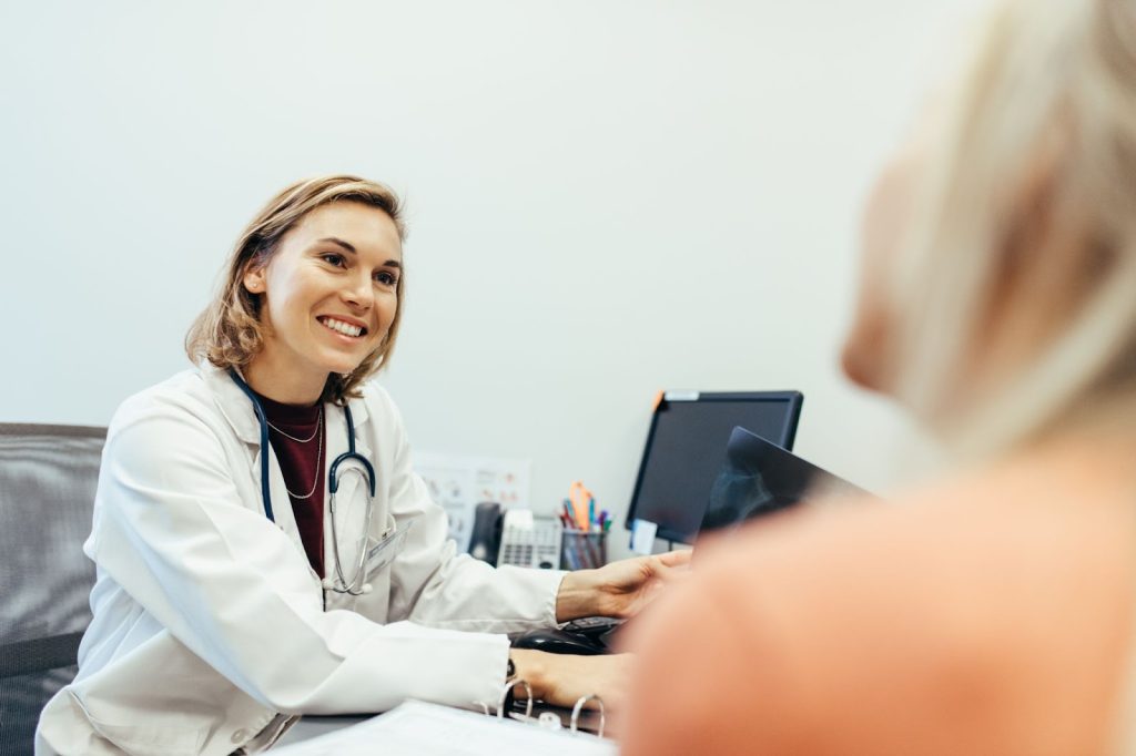 Médica branca sorrindo ao realizar anamnese com uma paciente. Ambas estão em consultório médico.