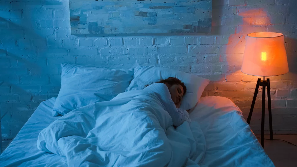 Mulher dormindo em uma cama com travesseiros e cobertor brancos. O quarto está em tons de azul e existe uma luminária com luz amarela acesa ao lado da cama.