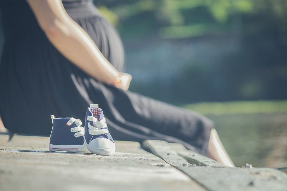 Imagem de sapatinhos de bebê. Como fundo, há uma mulher grávida sentada ao chão, de vestido preto e com as mãos sob a barriga.