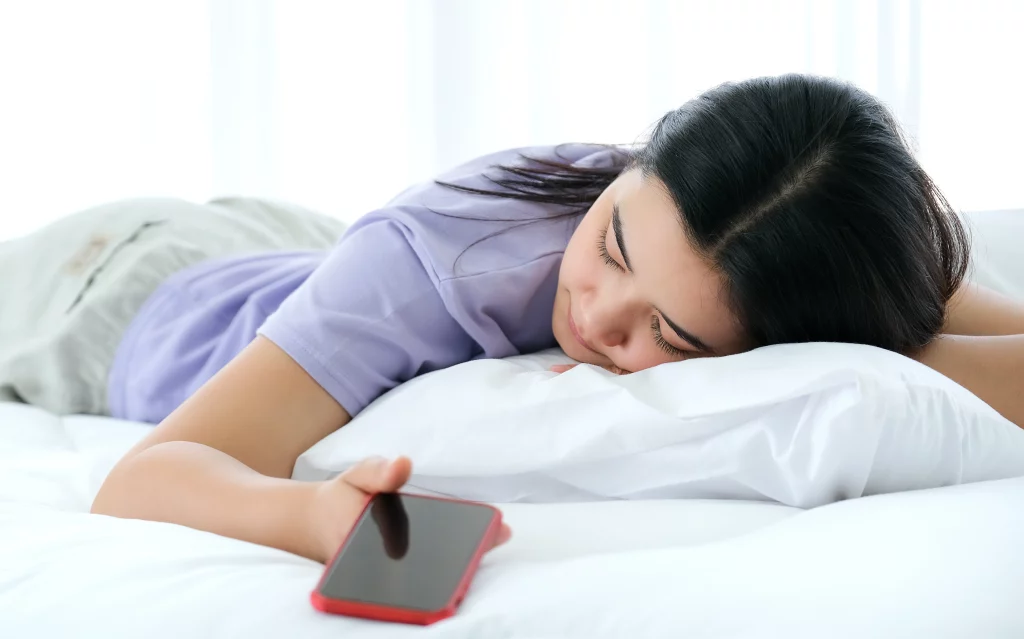 Mulher com camiseta roxa dormindo deitada de bruços em uma cama. Em sua mão há um smartphone de capa vermelha desligado.