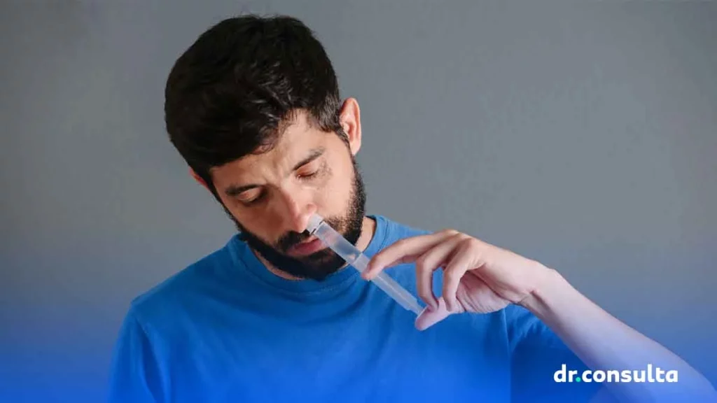 Homem de pele branca, cabelo e barba pretos e camiseta azul fazendo lavagem nasal com uma seringa.