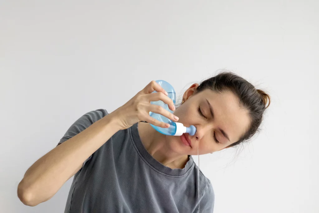 Mulher de pele branca e camiseta cinza com a cabeça inclinada ao realizar a lavagem nasal com um aparelho específico para o processo.