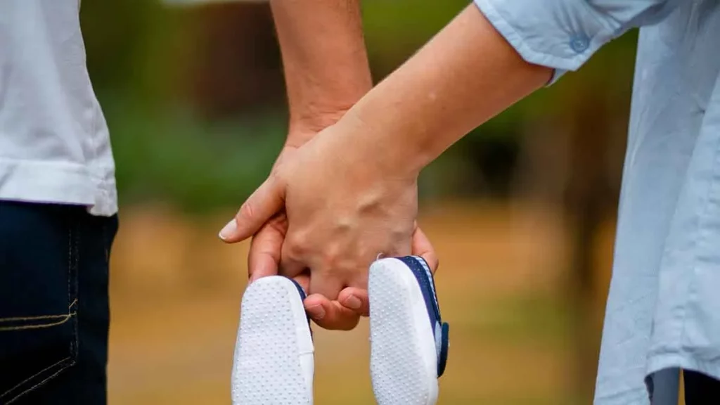 Casal de mãos dadas segurando um sapatinho de bebê. Na foto, seus rostos não aparecem.