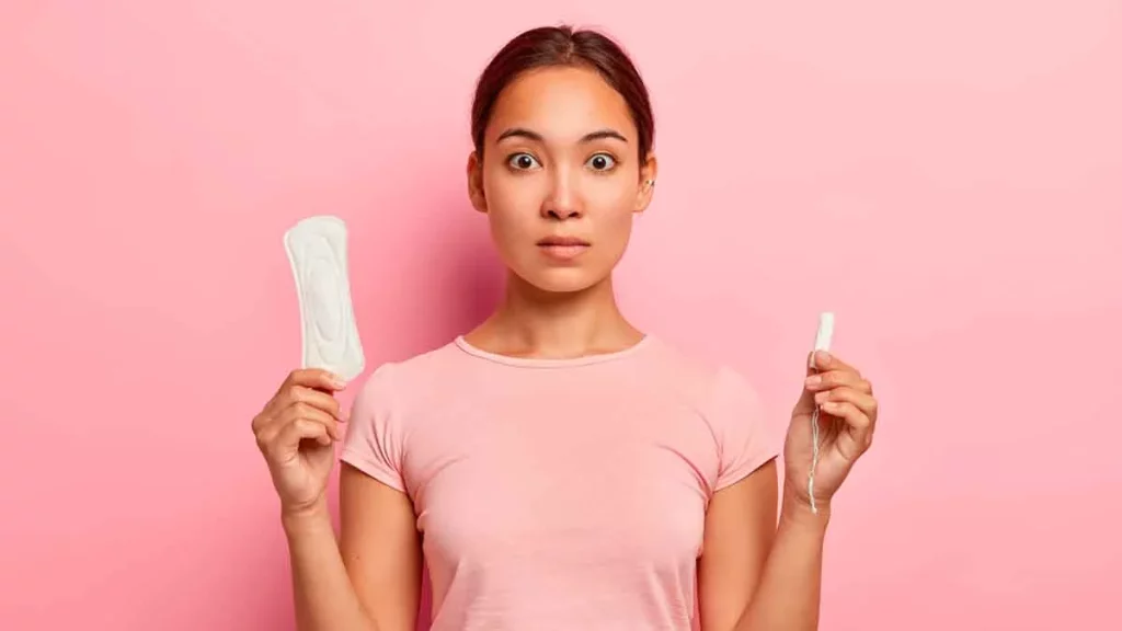 Mulher de camiseta rosa (combinando com a parede ao fundo) segurando um absorvente em cada mão.