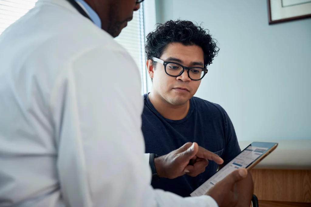 A foto mostra um médico negro conversando com paciente jovem branco de cabelos encaracolados e óculos enquanto mostra resultados de exames em um tablet. Apenas o rosto do paciente aparece.