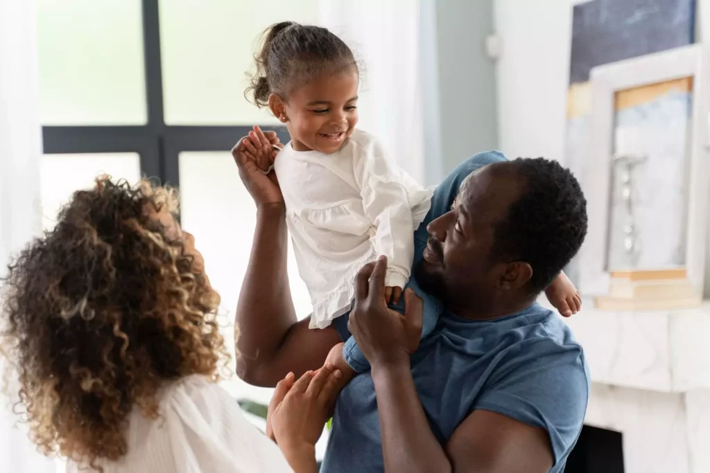 A imagem mostra uma família. Um homem de pele escura segura sua filha pequena nos seus ombros enquanto a mãe, na frente de ambos, os olha sorrindo.