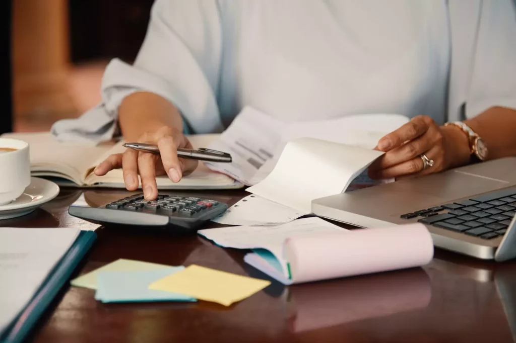 Vista de braços femininos fazendo contas em uma calculadora e com papéis coloridos. Também há um notebook e uma xícara sobre um pires (ambos brancos).