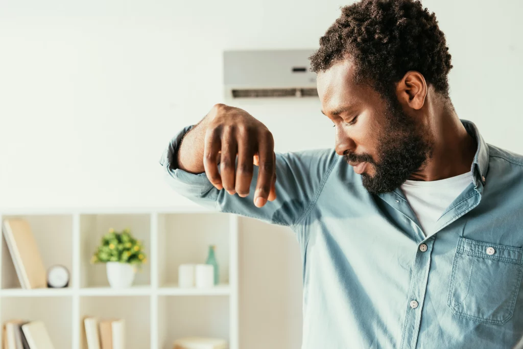 A imagem mostra um homem negro de camisa azul de botão. Ele está suando bastante e, ao fundo, existe uma estante com objetos e um aparelho de ar condicionado.