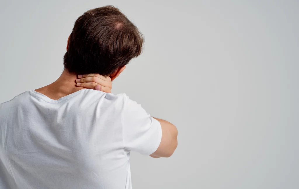 Na imagem é possível ver um homem branco de cabelos castanhos e camiseta branca. Sua cabeça está inclinada para o lado direito e uma de suas mãos está na região como se sentisse dor. Só é possível ver as costas do homem.