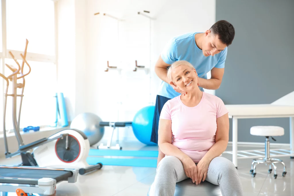 Na imagem há um fisioterapeuta cuidando do pescoço de uma paciente mulher, que está sentada à frente dele num espaço próprio para esse tipo de atendimento. Nesse espaço existem equipamentos fisioterapêuticos como esteiras de caminhada e corrida e bolas de pilates, além de maca e banco.