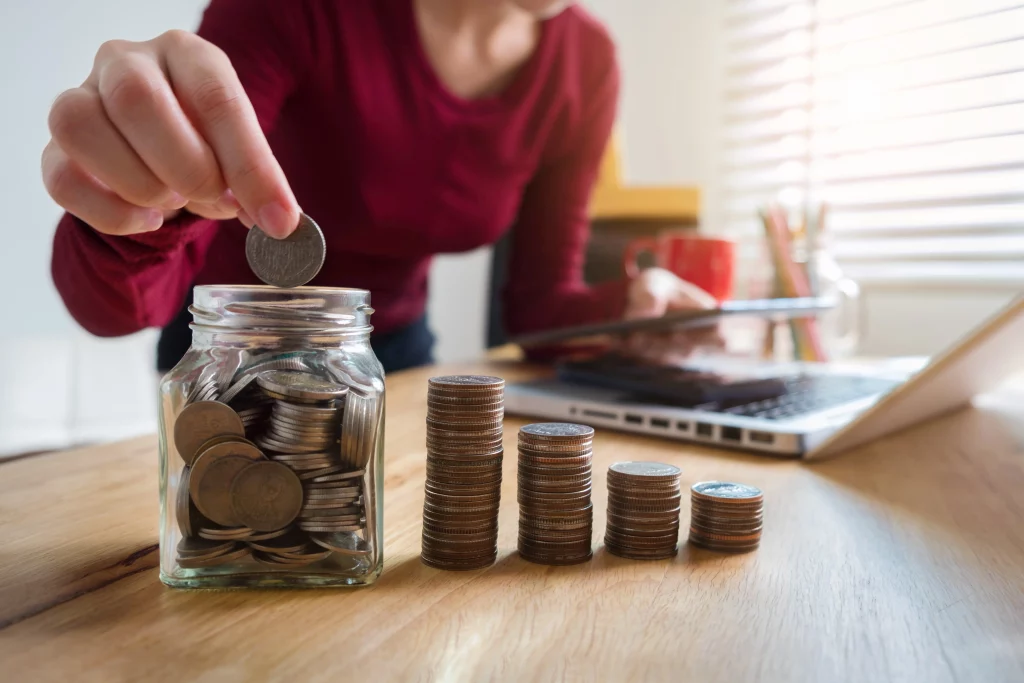 Na imagem é possível ver uma mulher colocando uma moeda em um recipiente de vidro que contém outras moedas, como se fosse um cofrinho. Ao lado do recipiente existem quatro pequenas pilhas de moedas formando uma escada. O rosto da mulher não aparece.