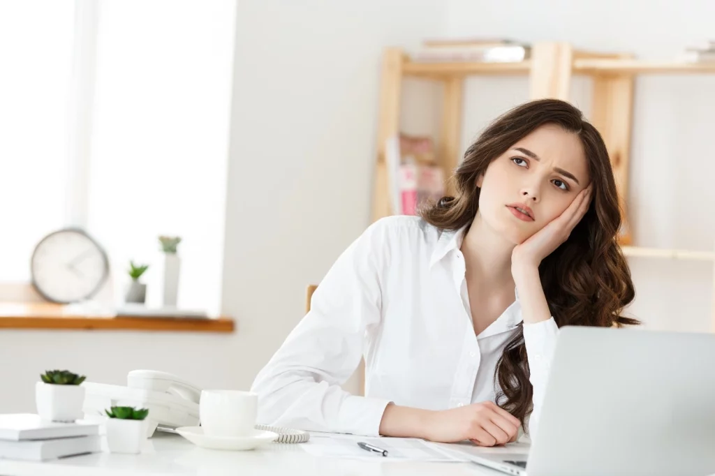 Mulher pensativa em uma mesa com notebook, xícara e papel. Ela veste uma camisa branca, combinando com o restante do ambiente todo branco.