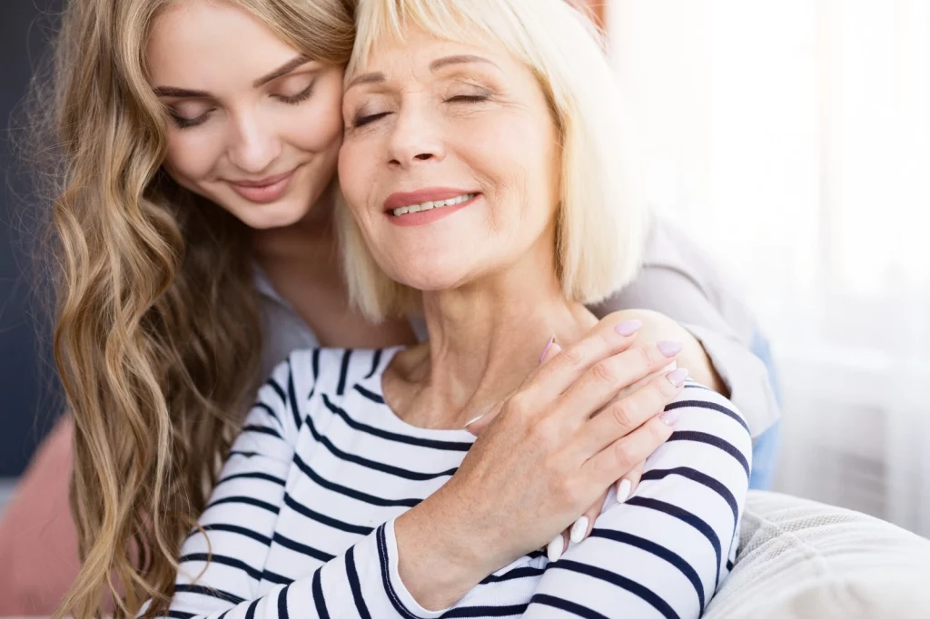 Na imagem existem duas mulheres, uma mais nova em pé e outra mais velha sentada. Ambas sorrindo e se abraçando de olhos fechados.