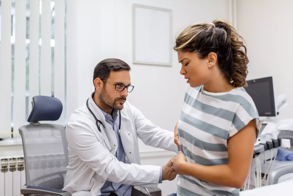 Na imagem é possível ver um médico branco de cabelos escuros e óculos realizando um exame físico no abdômen de uma paciente com camiseta listrada e cabelos presos. Ambos estão em um consultório onde há, também, um aparelho de ultrassonografia ao fundo.