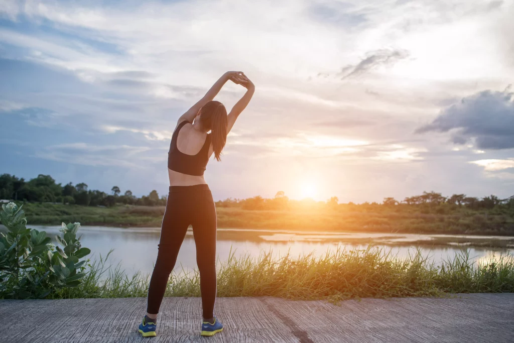 Na imagem é possível ver uma mulher de costas se alongando vestindo roupas pretas próprias para a realização de exercícios físicos. Ao fundo existe uma paisagem com céu azul, lago e vegetação.