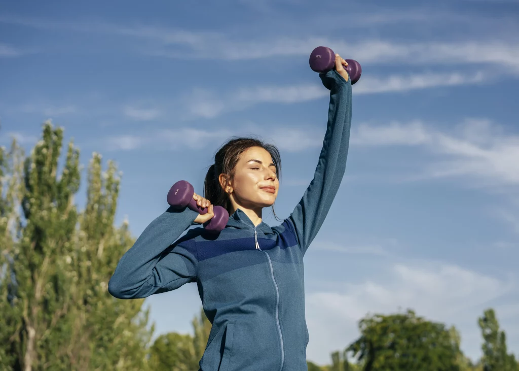 Na imagem é possível ver uma mulher de meia idade fazendo exercícios físicos ao ar livre. Ela está carregando halteres roxos e está de blusa de frio azul.