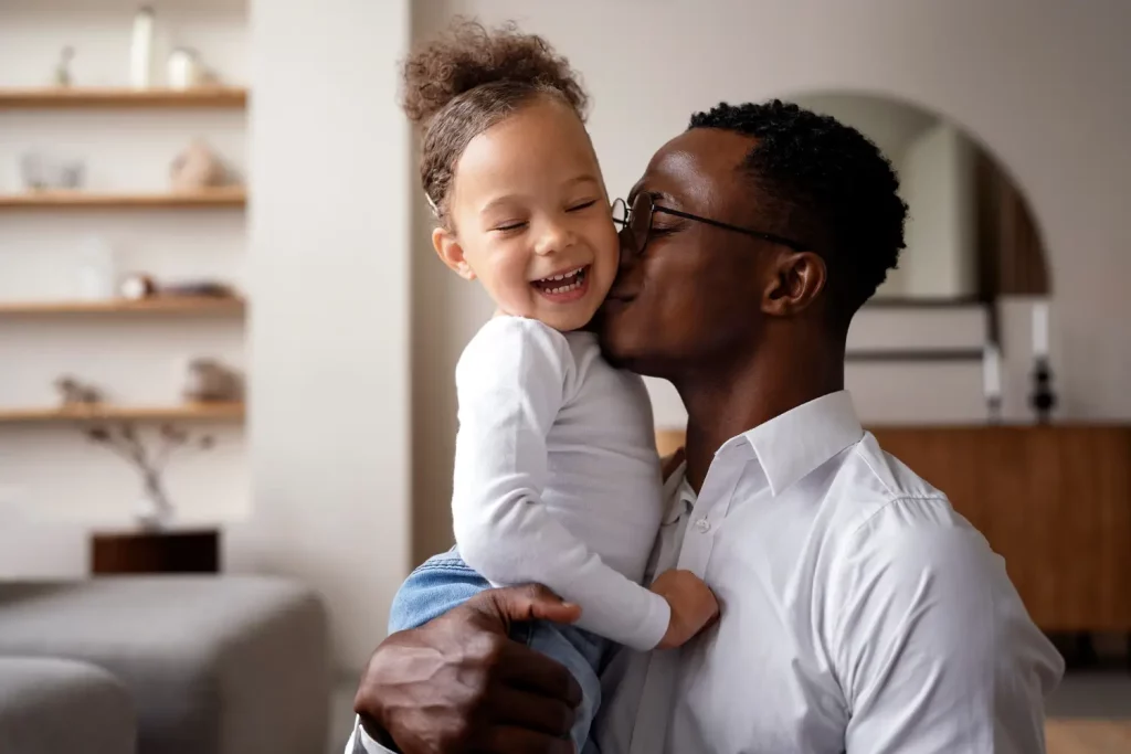 Na imagem é possível ver um pai negro abraçando sua filha pequena. Ela tem a pele mais clara, está sorrindo e, assim como ele, ela está de olhos fechados. Ambos vestem roupas brancas e têm cabelos escuros.