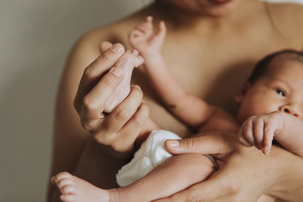 Mulher segurando um bebê recém-nascido junto ao seu corpo. Enquanto o segura, ela também está tocando os pés do bebê com as pontas dos dedos.