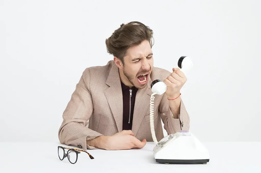 Um homem branco, vestindo um terno marrom, grita em uma ligação telefônica expressando raiva intensa.