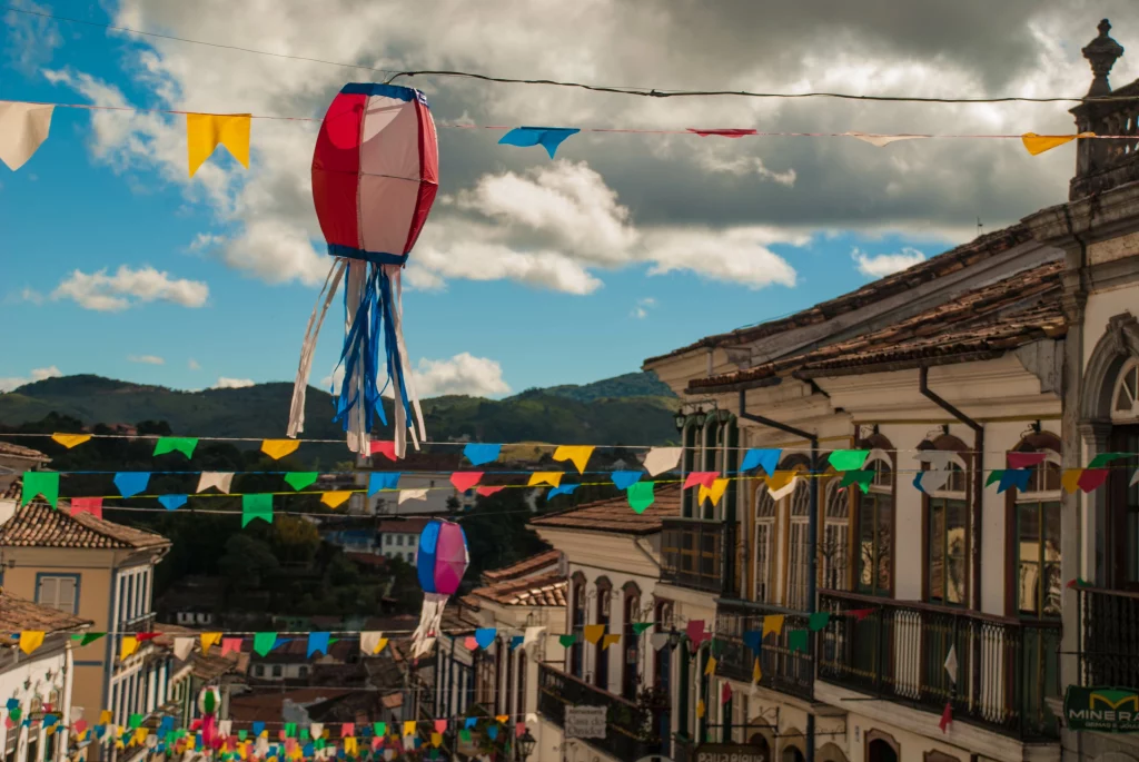 Na imagem, há bandeiras de papel coloridas penduradas em casas formando uma decoração de festa junina.