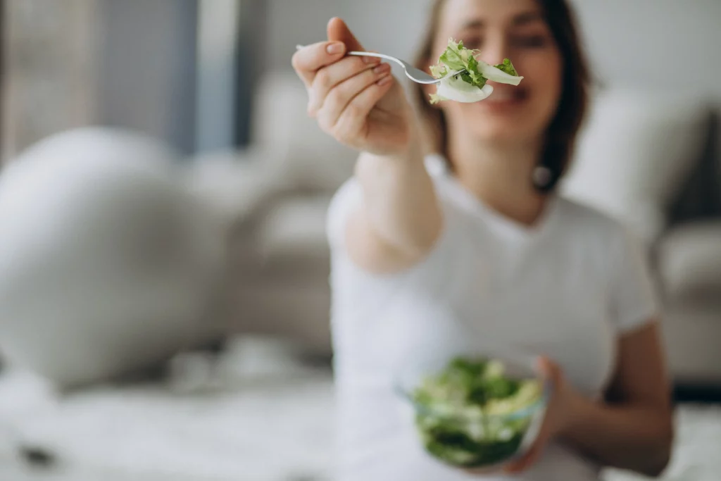 Na imagem é possível ver uma mulher vestindo roupas brancas e com o braço estendido para a câmera, oferecendo um garfo com verduras.