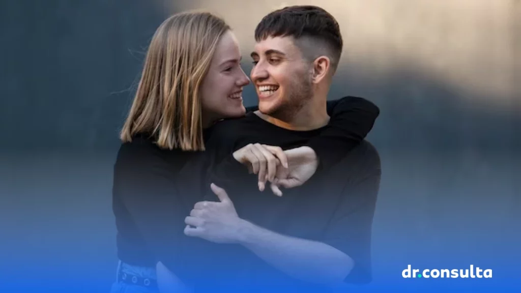 Na imagem é possível ver um casal sorrindo. Ambos estão de roupas pretas e se olham ao mesmo tempo que sorriem. Uma das pessoas é um homem trans.