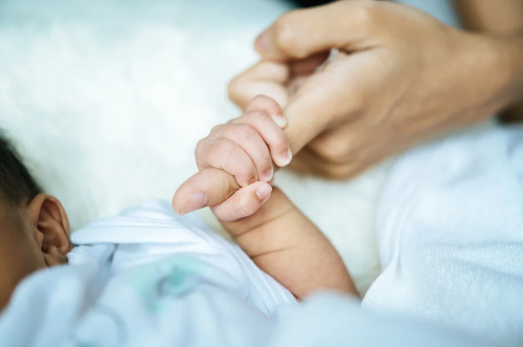 A imagem apresenta um bebê segurando o dedo indicador de sua mãe. Na foto, o rosto da criança não aparece. 