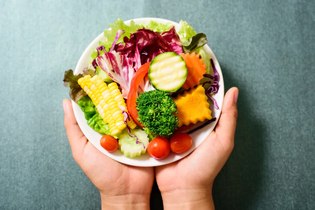 Imagem de duas mão segurando um prato branco que contém alimentos considerados saudáveis como legumes e vegetais.