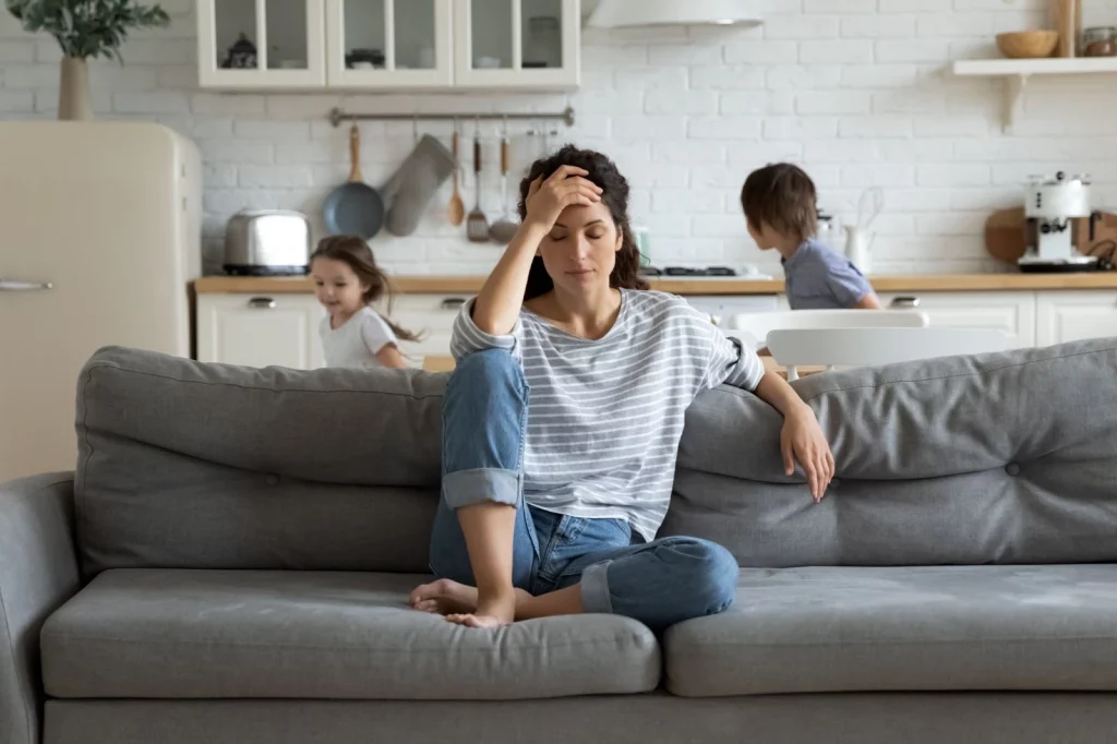 A imagem apresenta uma mãe cansada e estressada enquanto crianças correm ao redor da sala.