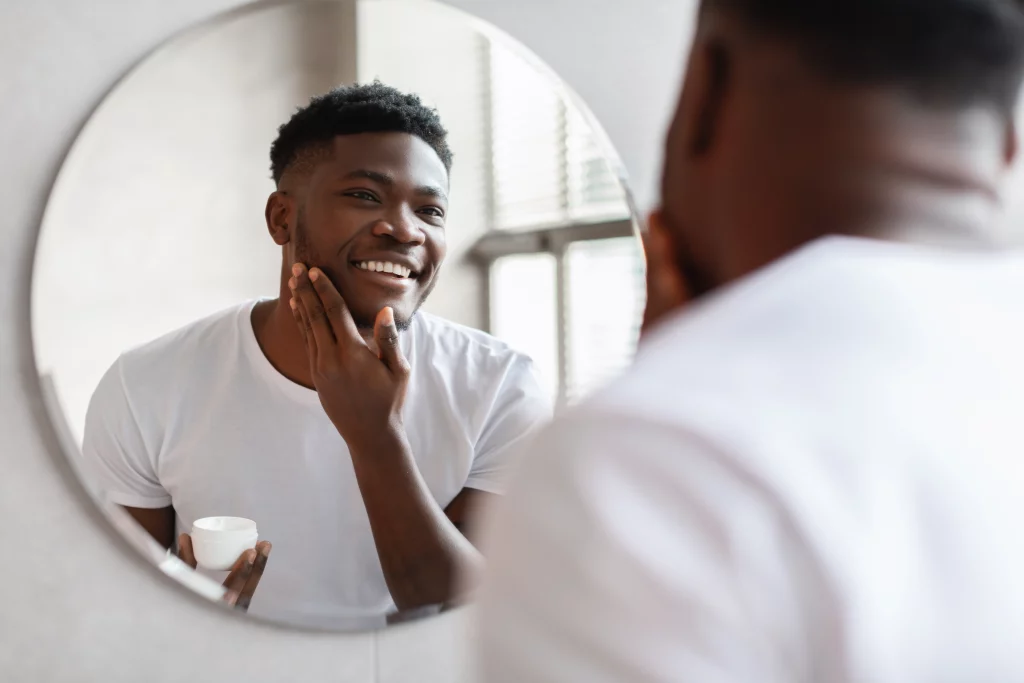 A imagem apresenta o reflexo de um homem negro cuidando da pele facial em um espelho redondo. 