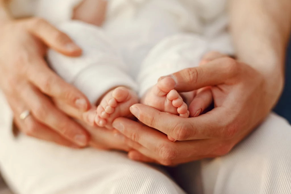 A imagem apresenta uma mãe segurando seu bebê no colo. Ela está envolvendo os pés da criança em suas mãos. O rosto da mãe e criança não aparecem na imagem.