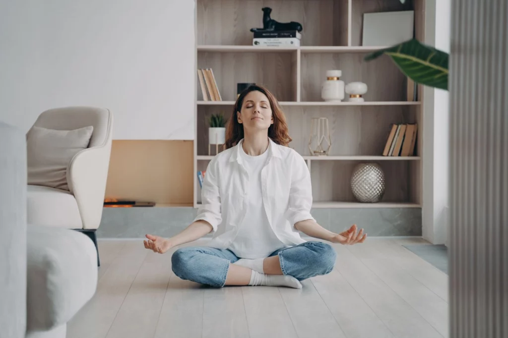 Imagem de uma mulher branca vestindo uma blusa branca e calça jeans. Ela está em posição de meditação.