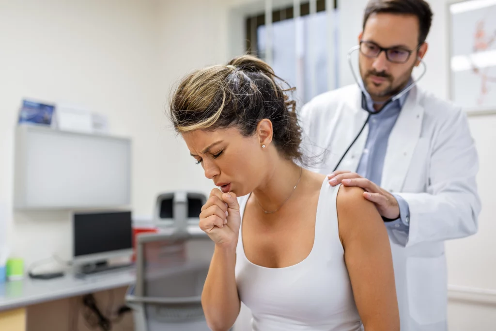 Imagem de um médico examinando as costas de uma paciente tossindo ao utilizar um estetoscópio.