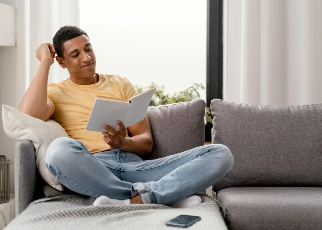 Imagem de um homem negro sentado no sofá lendo um livro.