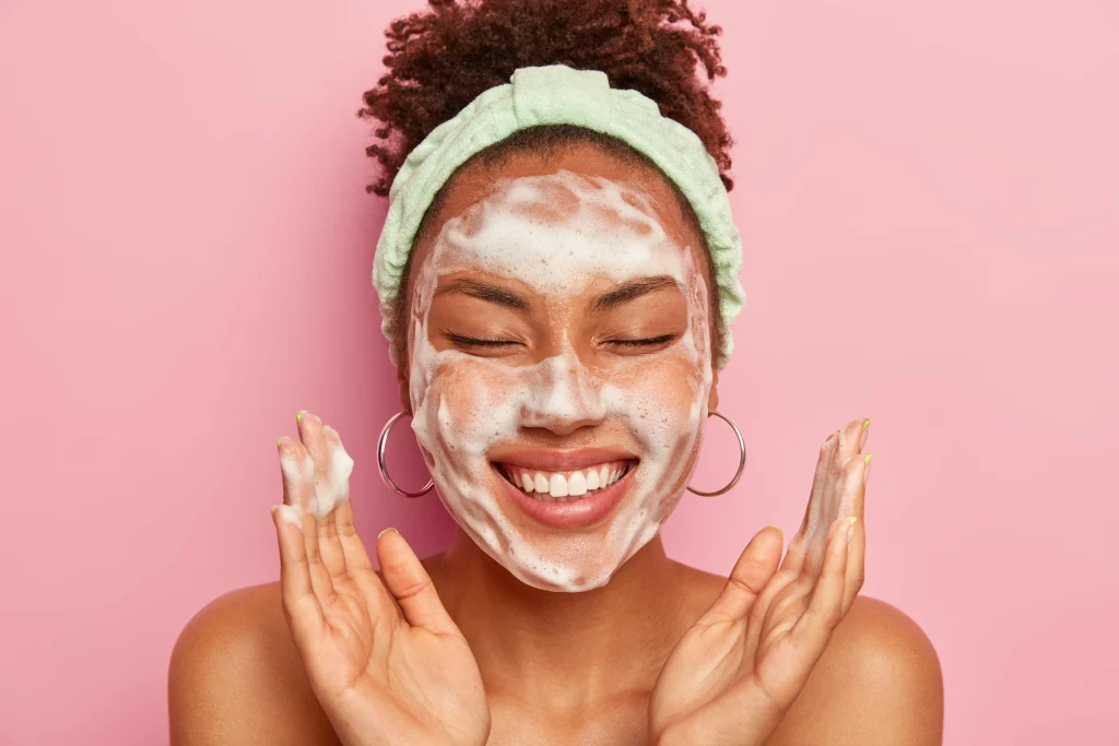 Imagem de uma mulher negra sorrindo de olhos fechados e com espuma no rosto como se o estivesse lavando.