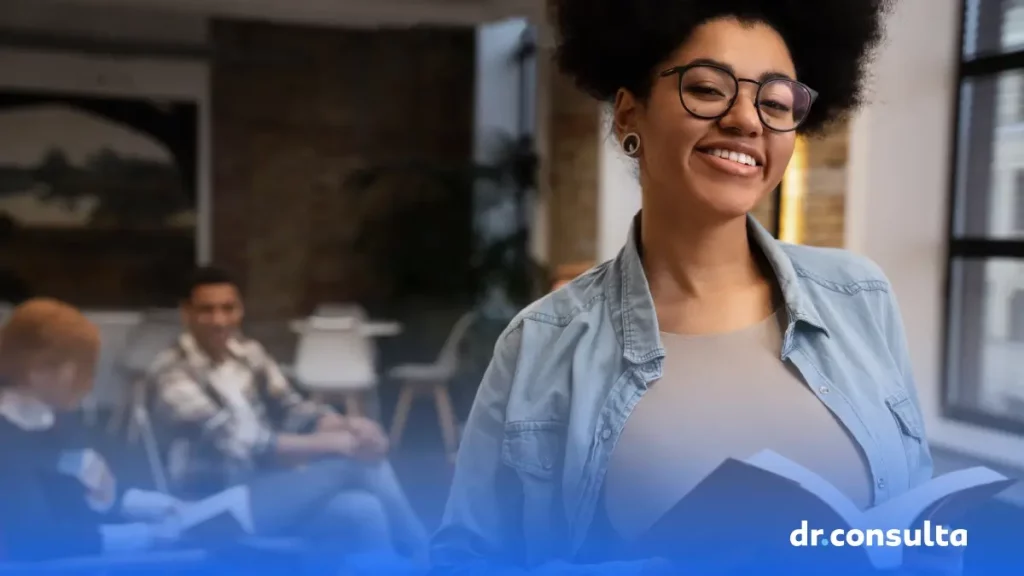 Imagem de uma jovem de camisa bege e jaqueta jeans segurando um livro. Ela está posando para a câmera e sorri.