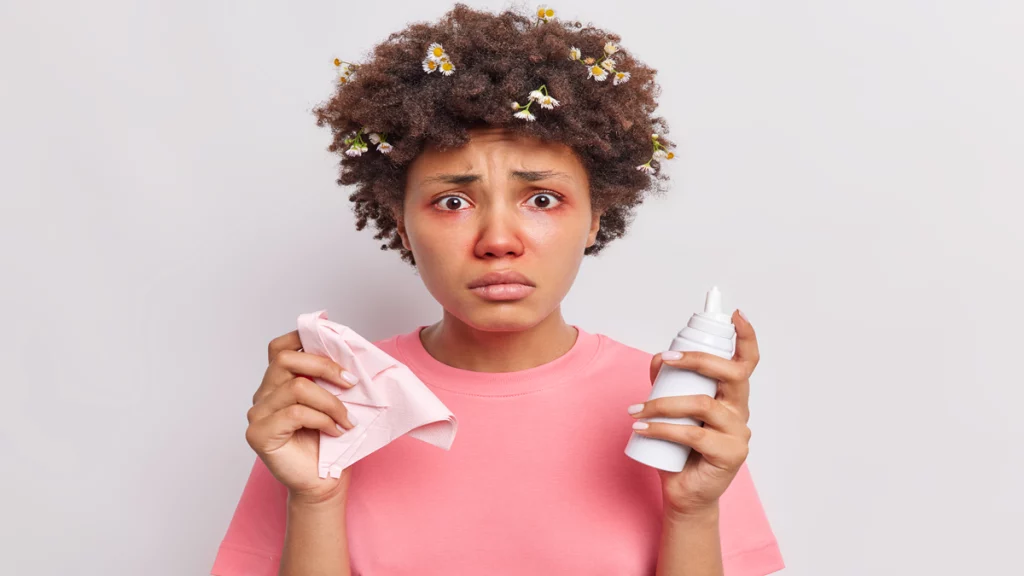 Imagem de uma mulher com os olhos e nariz vermelhos durante uma reação alérgica. Além disso, ela segura um lenço de pano e um soro fisiológico nas mãos.